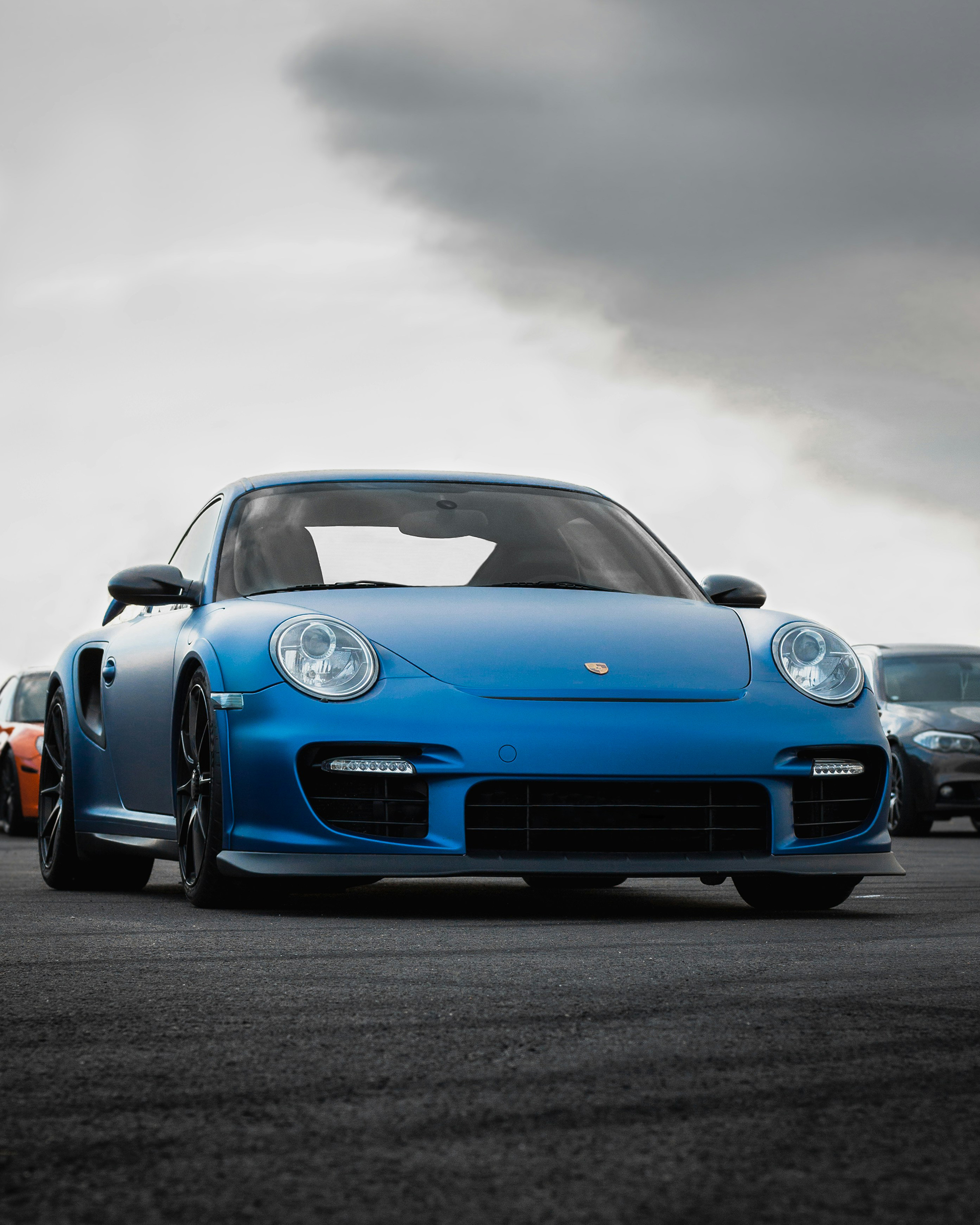 Front picture of a light blue porsche 911, with the background having two cars on the left and on the right